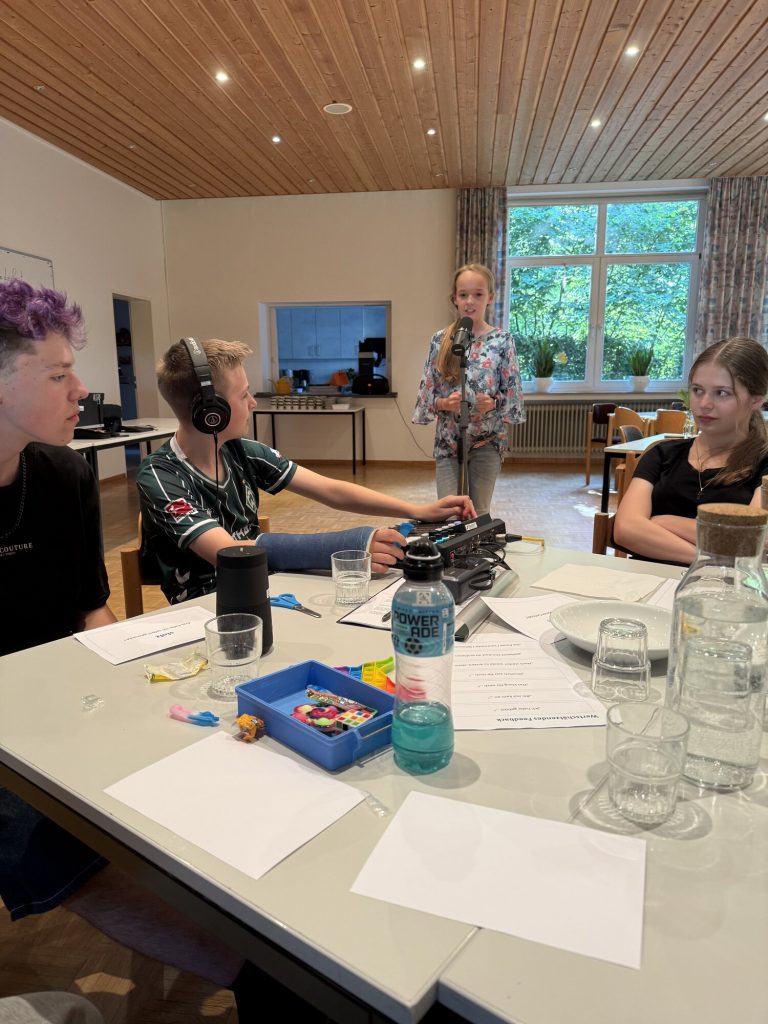 Vier Konfis arbeiten an einem Mikrofon und einem Audiomischpult; eine Jugendliche spricht im Stehen.