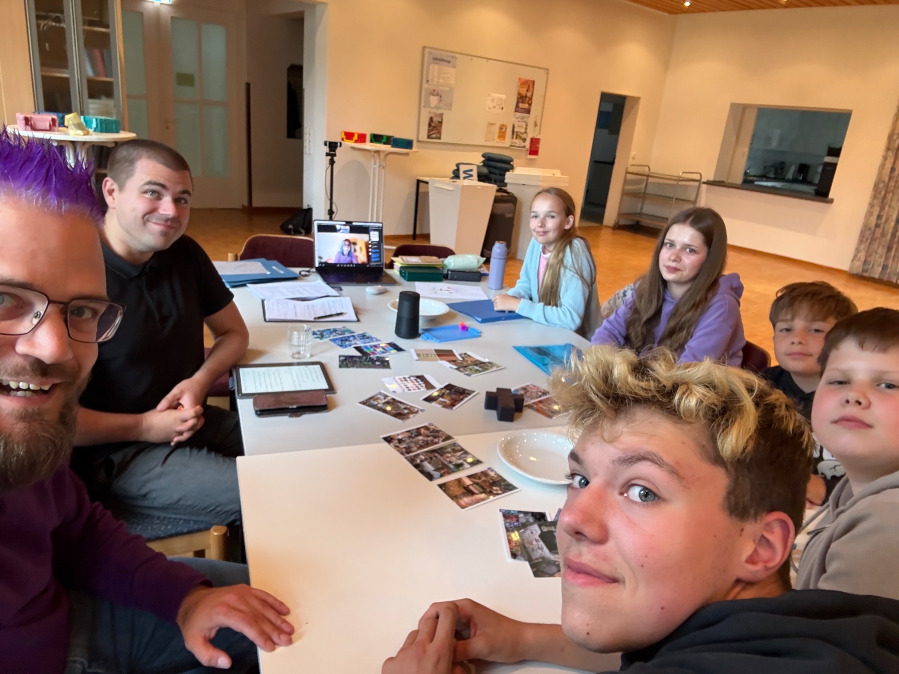 Christoph und Lars sitzen mit mehreren Konfis am Werkstatttisch; Amy ist auf einem Laptopbildschirm zugeschaltet.