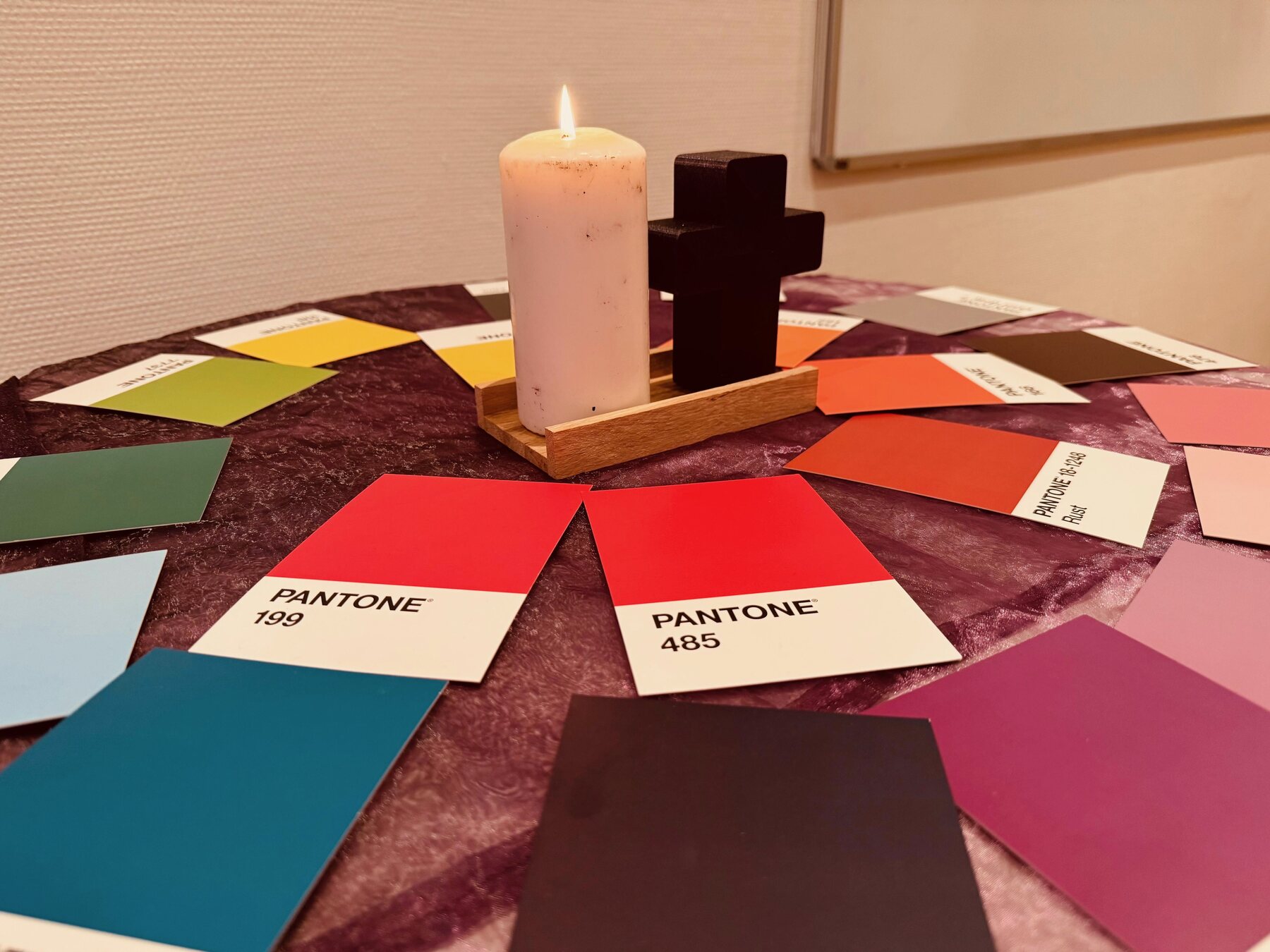Eine brennende Kerze und ein Kreuz stehen zwischen bunten Farbkarten auf einem violetten Tuch.